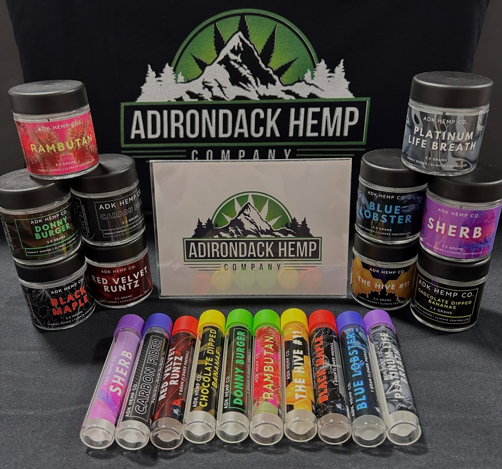 ADK Hemp Co. branded table display at a 21+ pop-up event — jars of small-batch hemp flower (Rambutan, Black Maple, Red Velvet Runtz, Double Burger, Platinum Life Breath, Blue Lobster, Sherb) and a row of pre-roll tubes arranged around the Adirondack Hemp Company logo.